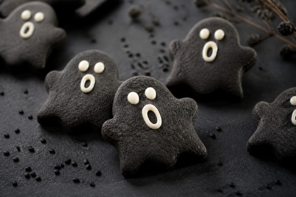 Black sesame ghost cut-out cookies with white chocolate face dots on a dark Halloween table
