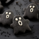 Black sesame ghost cut-out cookies with white chocolate face dots on a dark Halloween table