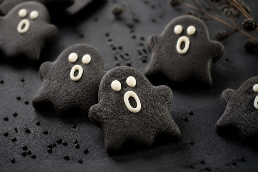 Black sesame ghost cut-out cookies with white chocolate face dots on a dark Halloween table