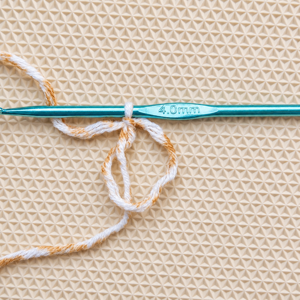 Magic circle formed with ivory and tan cotton yarn beside a teal 4 mm crochet hook.